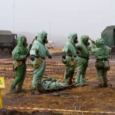 Żołnierze w kombinezonach ochronnych, gotowi na zagrożenie iperytem, ćwiczą ewakuację rannego.