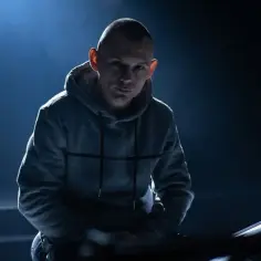 Młody polski raper w szarej bluzie z kapturem, oświetlony od tyłu, zamyślony.