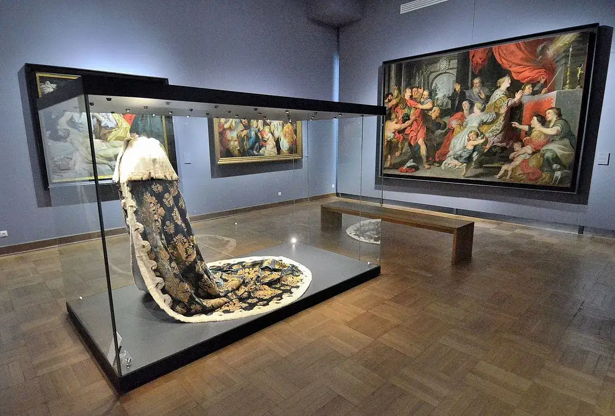 W muzeum kustosz podziwia bogato zdobiony strój królewski w gablocie, obok wiszą wielkie obrazy.